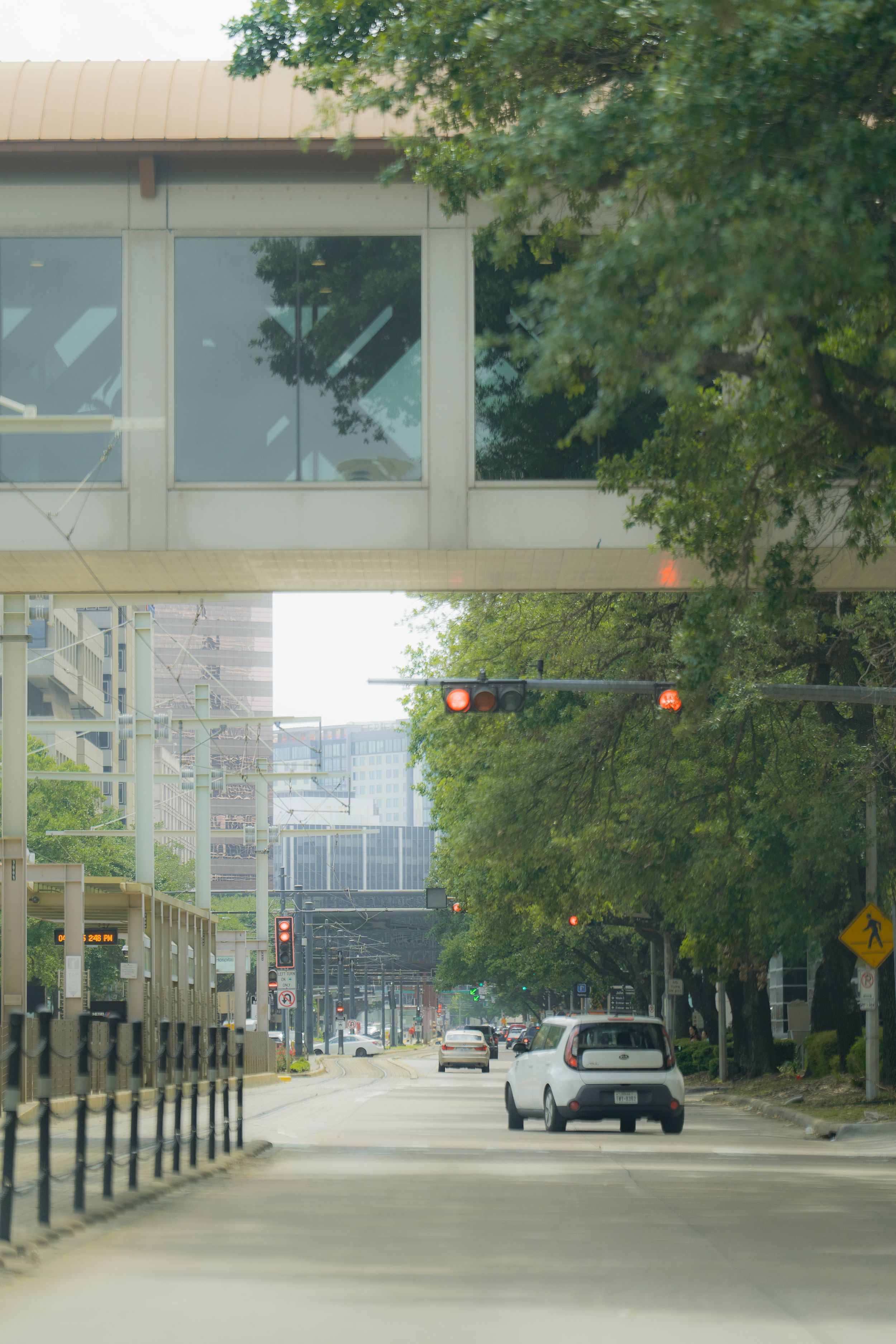 Urban street with pedestrian skybridge and tall buildings bordered by trees and traffic lights in a downtown setting.