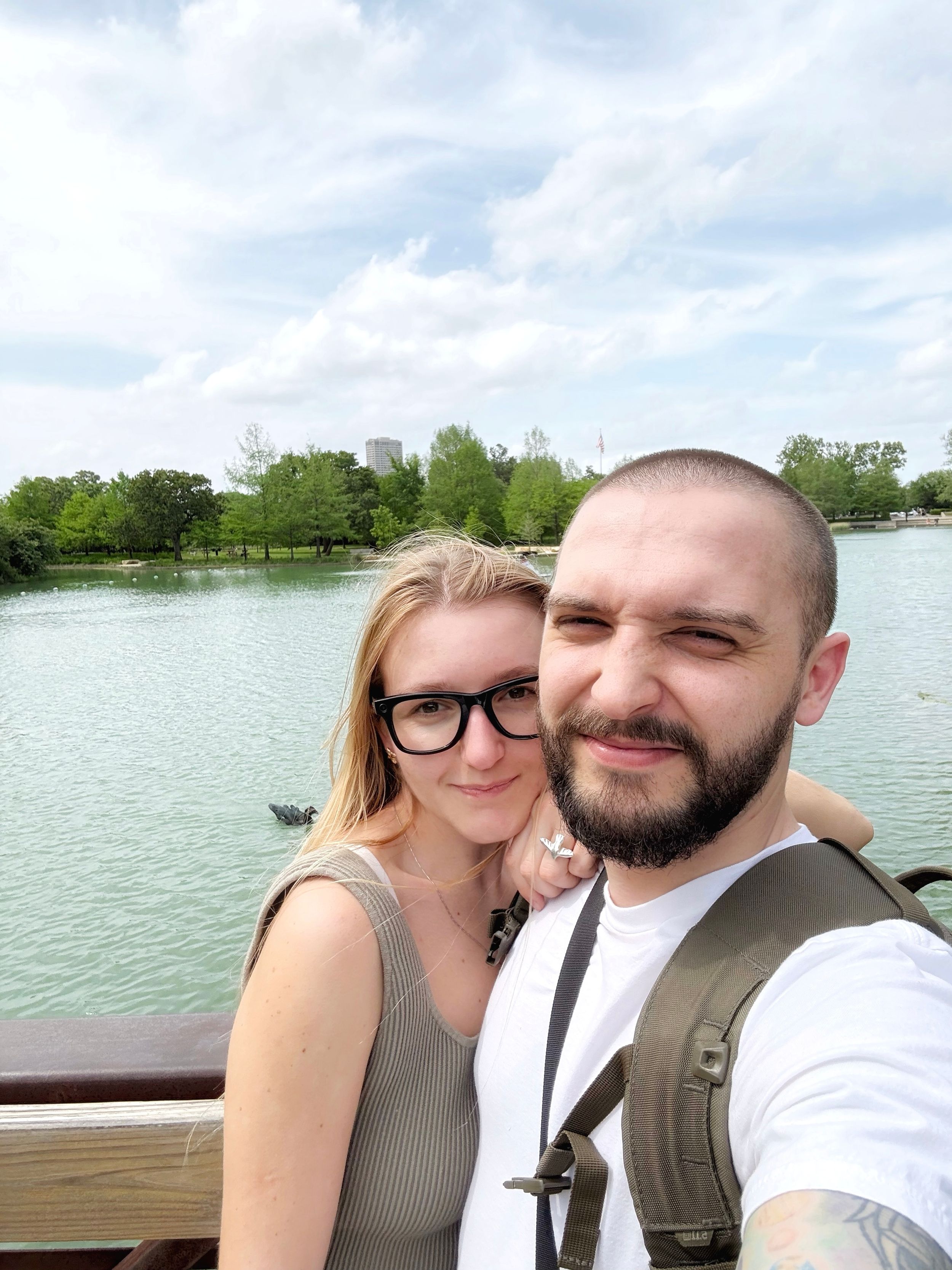A couple takes a selfie together on a wooden deck overlooking a serene lake with trees in the background.