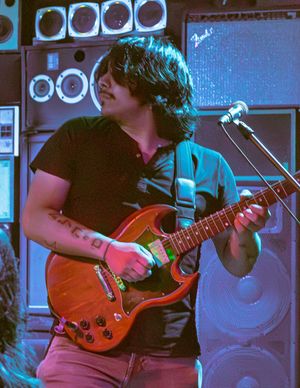 Musician playing an electric guitar on stage with dramatic blue lighting and musical equipment in the background.