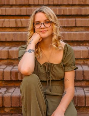 Portrait against brick stairs featuring someone in an olive green dress with glasses and blonde wavy hair.