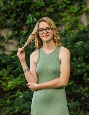 Someone wearing glasses and a sage green ribbed sleeveless dress poses outdoors with greenery in the background.