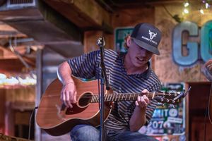 Musician playing acoustic guitar on stage at a bar, wearing a striped shirt and black cap.