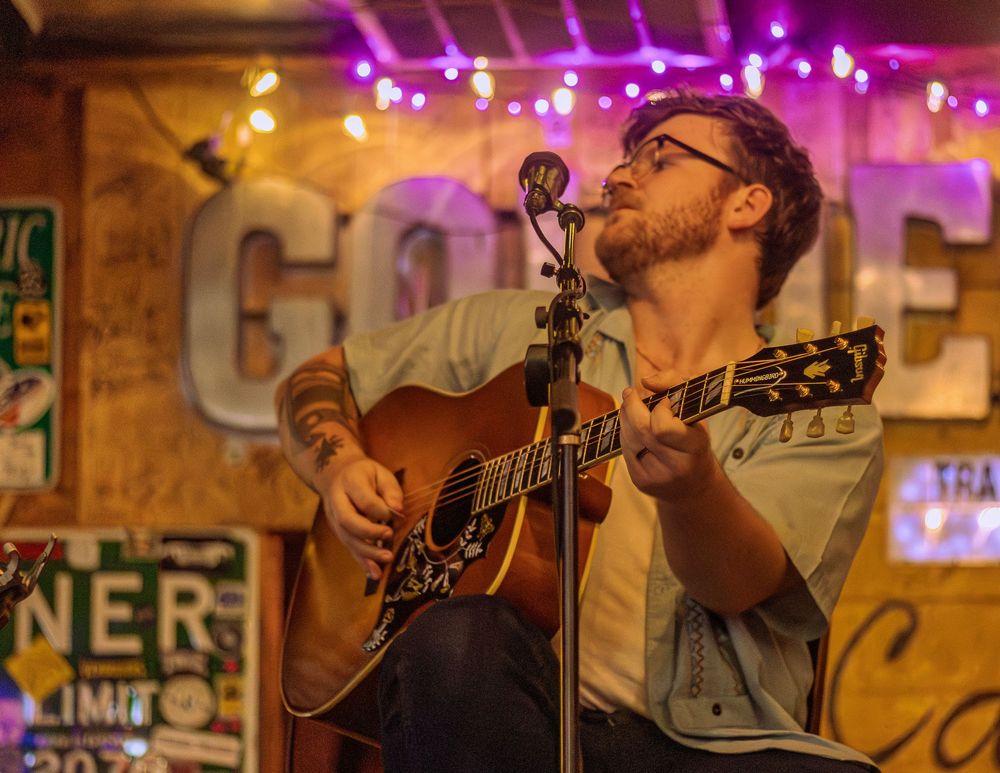 A musician performs with an acoustic guitar under purple stage lights at a small venue with neon signs in the background.