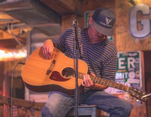 A musician in a striped shirt and baseball cap performs with an acoustic guitar on stage under warm lighting.