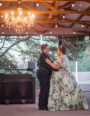 Couple shares first dance under chandelier and string lights in rustic pavilion, woman in floral ballgown.