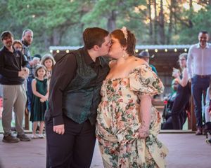Couple sharing first kiss at outdoor wedding reception with guests applauding in wooded venue.