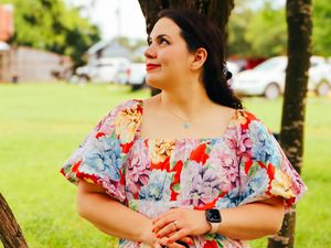 A person in a colorful floral dress with flutter sleeves smiles while standing in a grassy outdoor area.