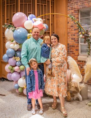A family poses together by a decorative balloon arch and brick wall with their golden retriever dog.