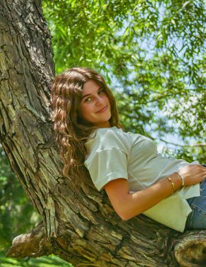 A young person in a white t-shirt reclines against a large tree trunk in a sunny outdoor setting with green foliage.
