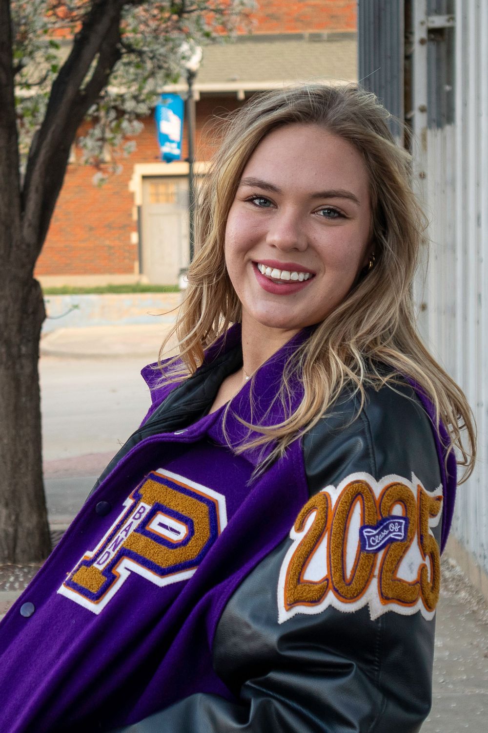 A student wearing a purple and black varsity letterman jacket poses outdoors near a brick building.