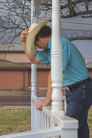 Person in blue shirt adjusting cowboy hat while leaning on white porch column.