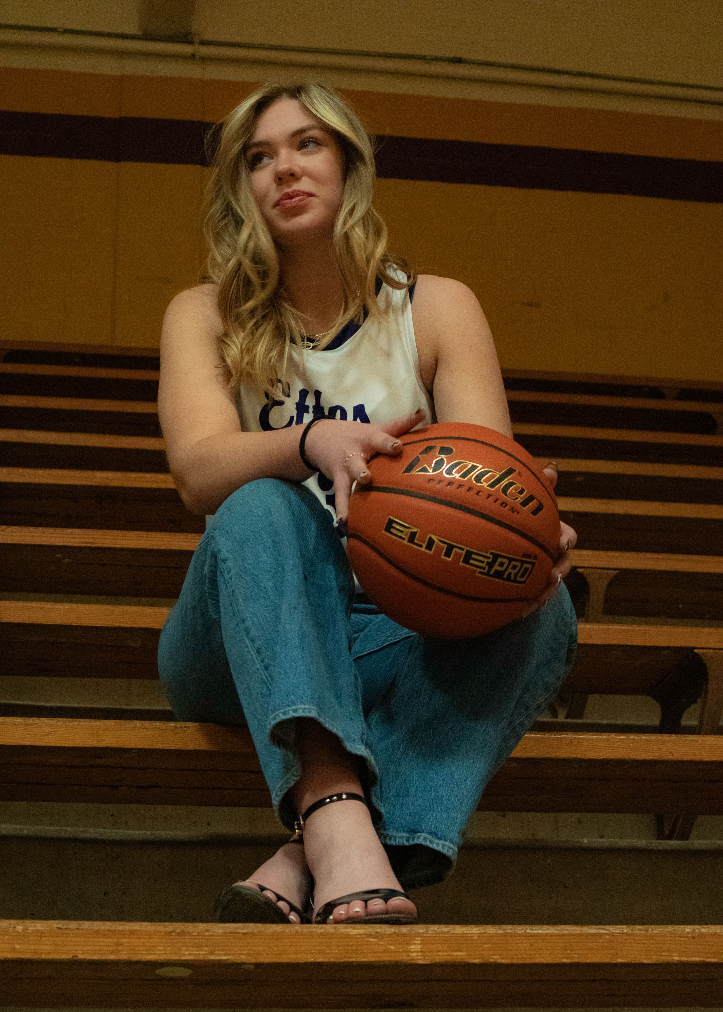 A seated figure in jeans and tank top holds a basketball while resting on wooden bleachers in a gymnasium.