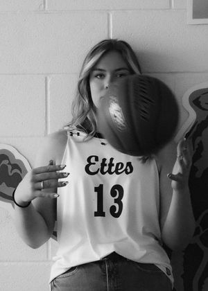 Basketball player wearing Ettes jersey number 13 poses with ball against white wall.