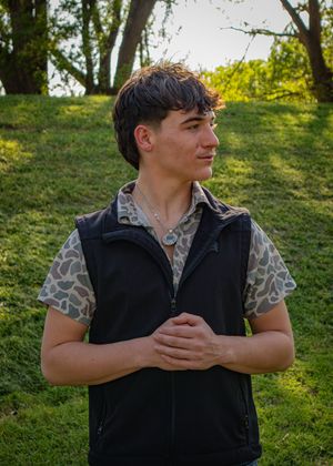 Person wearing a patterned shirt and black vest poses outdoors against a sunny green background.