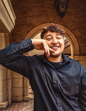 A person in a navy button-down shirt smiles joyfully while posing against a brick archway background.