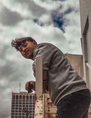 Looking up at someone in a gray sweatshirt leaning over a building edge against a cloudy sky backdrop in an urban setting.