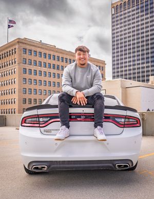 Someone in a gray sweater sits on the trunk of a white Dodge Charger in a downtown parking area with buildings in background.