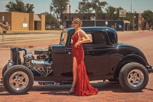 A vintage hot rod coupe parked on a brick street with someone in a flowing red formal gown standing beside it.