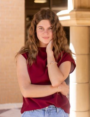 Young person posing in a burgundy sleeveless top and denim jeans against a beige stone column background.