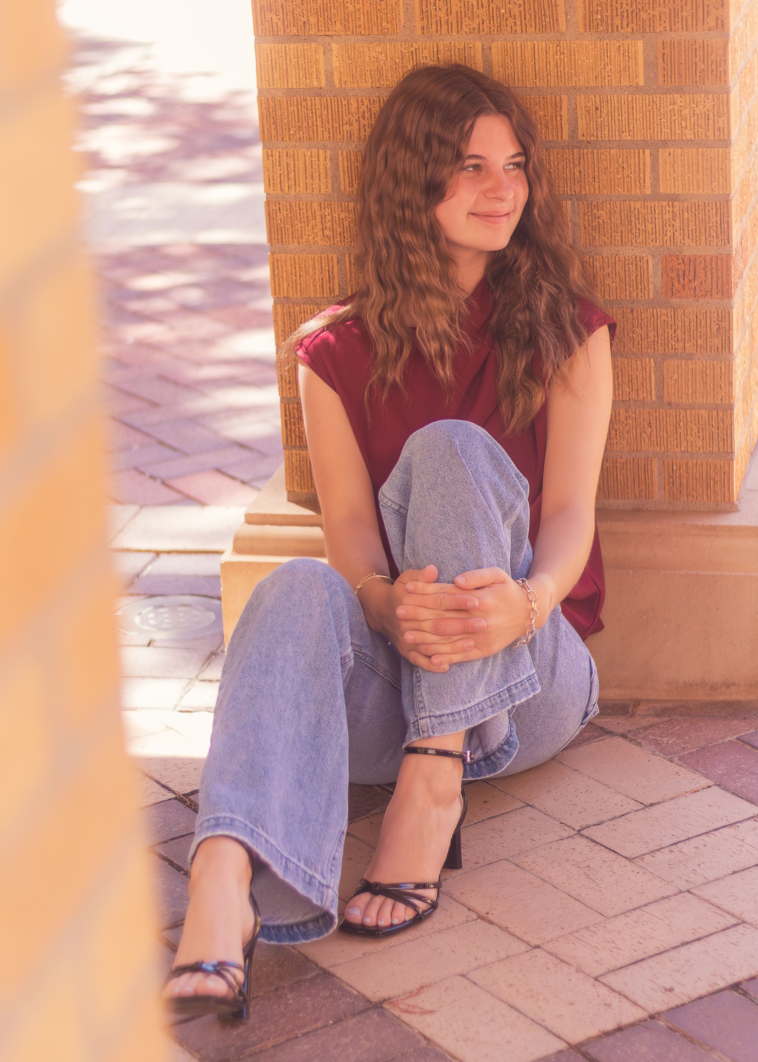 Someone in blue jeans and a burgundy sleeveless top sits on tiled steps in front of a brick wall.