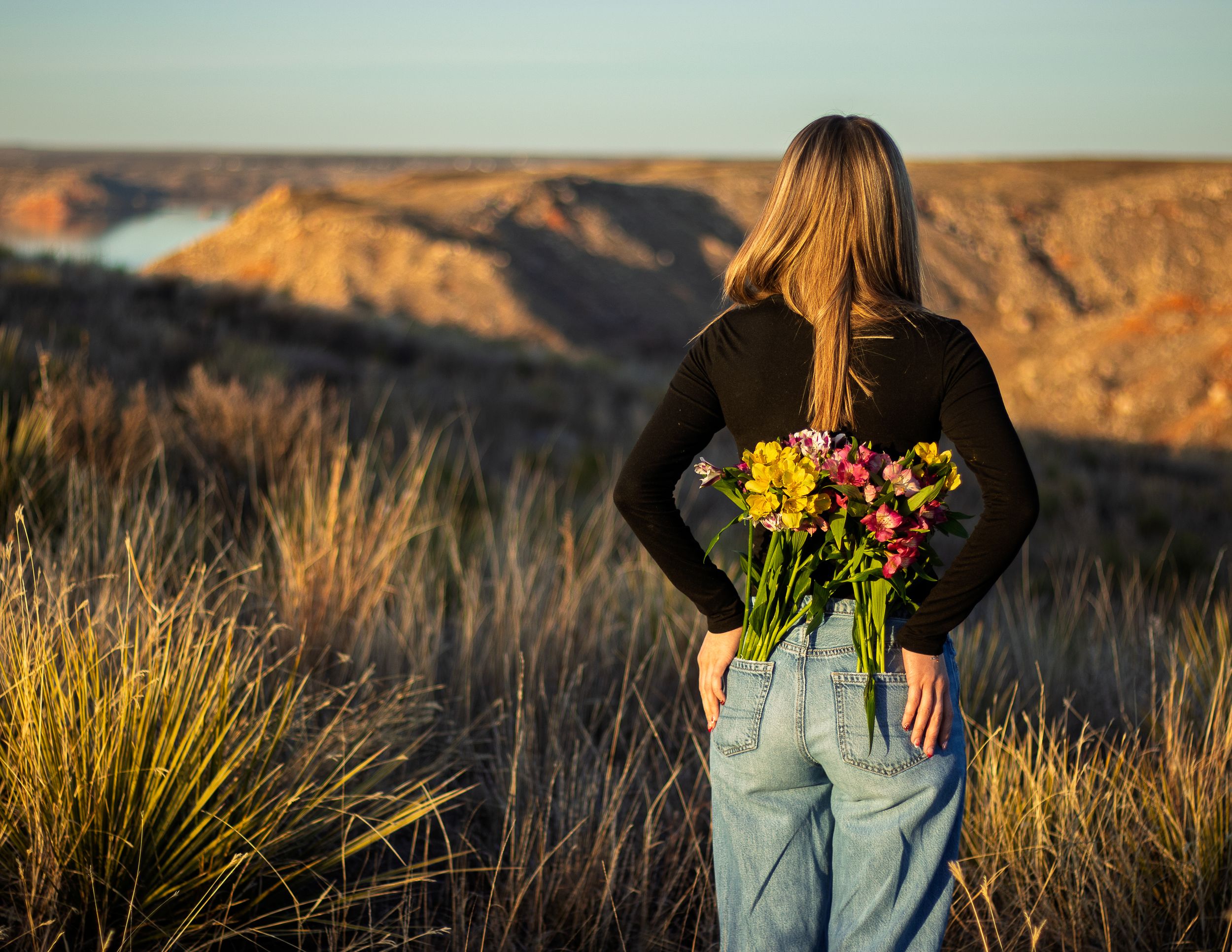 Woman holding colorful bouquet of flowers behind her back overlooking a scenic canyon landscape at golden hour.