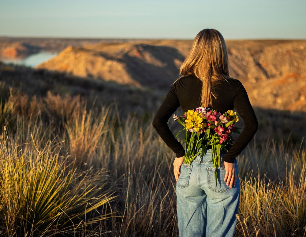 Woman holding colorful bouquet of flowers behind her back overlooking a scenic canyon landscape at golden hour.