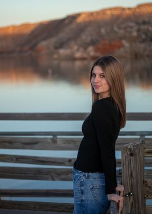 Young woman in black top and jeans smiling on a wooden dock with a serene lake and mountain backdrop at sunset.