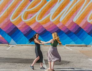 Two people dance joyfully in front of a vibrant mural featuring blue, pink, and orange geometric patterns.