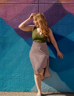 A woman in a stylish olive green top and grey skirt poses dramatically against a colorful gradient wall outdoors.
