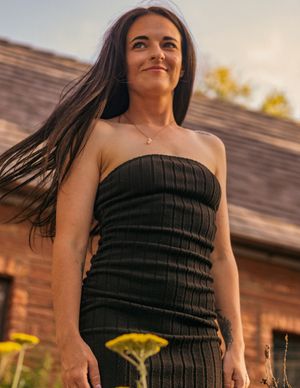 A smiling woman in a black strapless dress stands outdoors at sunset with wildflowers in bloom.