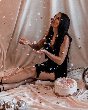 Woman in black clothing sits with white confetti falling around her in a pink-tinted indoor photoshoot setting.