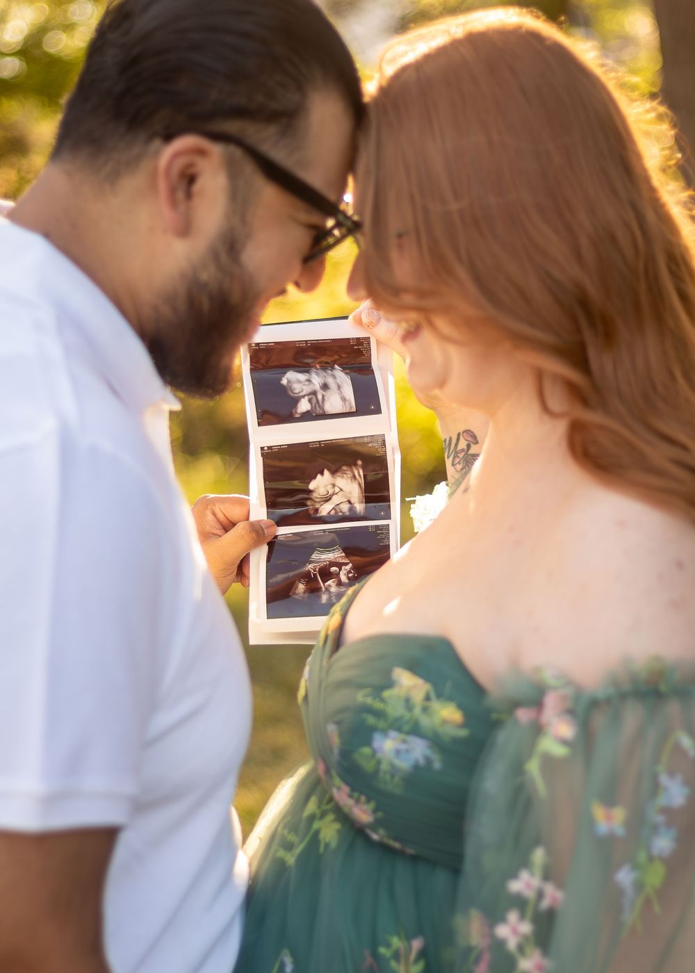 A couple in white and green attire share a tender moment while looking at ultrasound photos in a sunny outdoor setting.