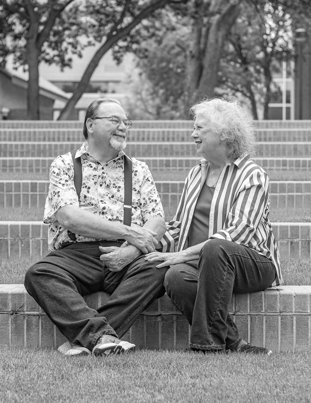 Elderly couple sitting on brick steps outdoors, laughing and holding hands in a black and white photo.