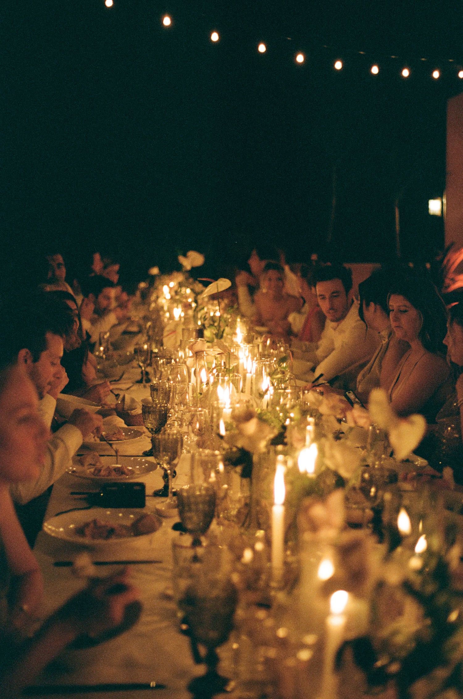An intimate candlelit dinner party with guests gathered around a long table under string lights.