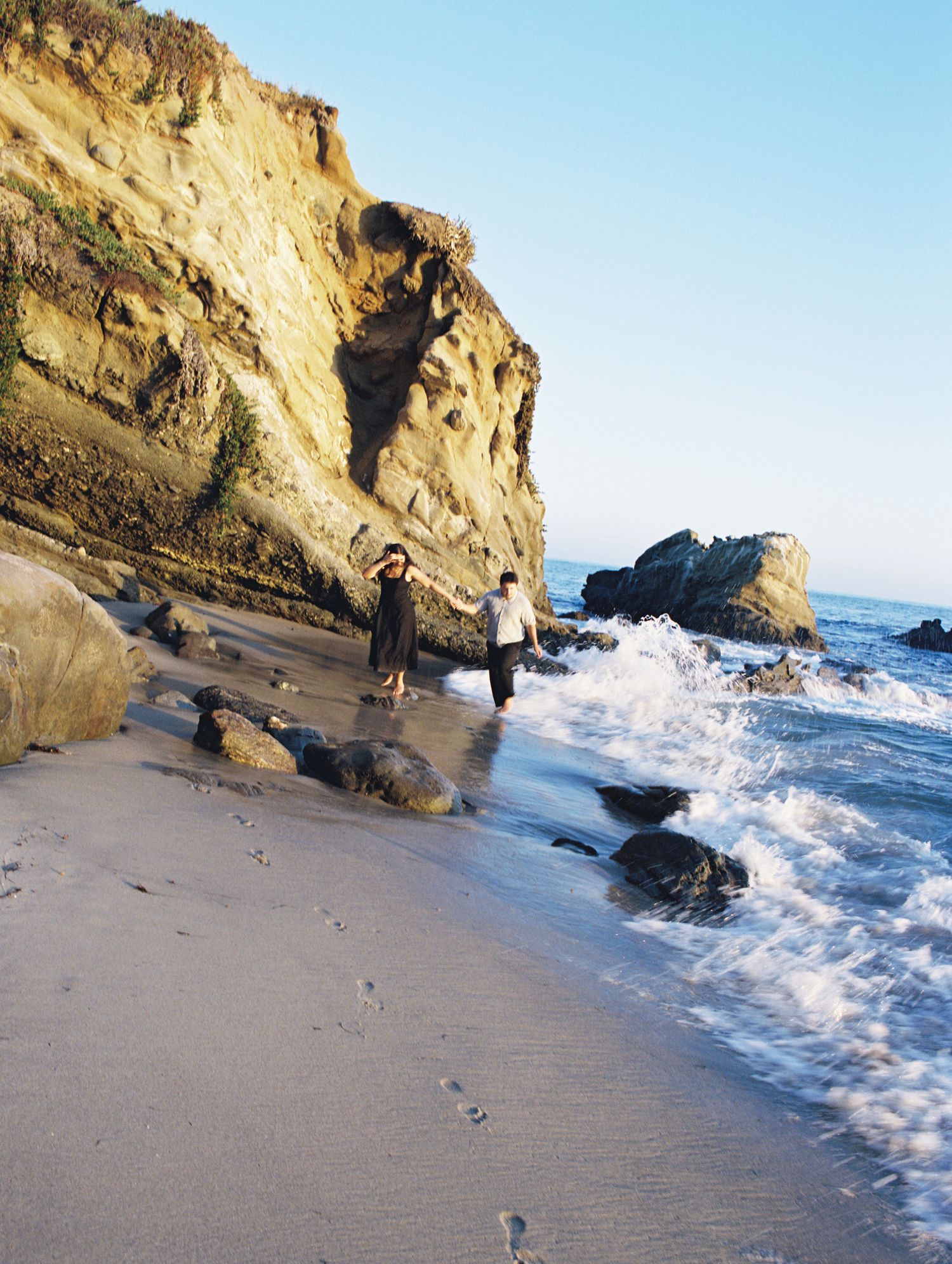 Two figures explore a scenic beach with dramatic cliffs and crashing waves along a rocky coastline.