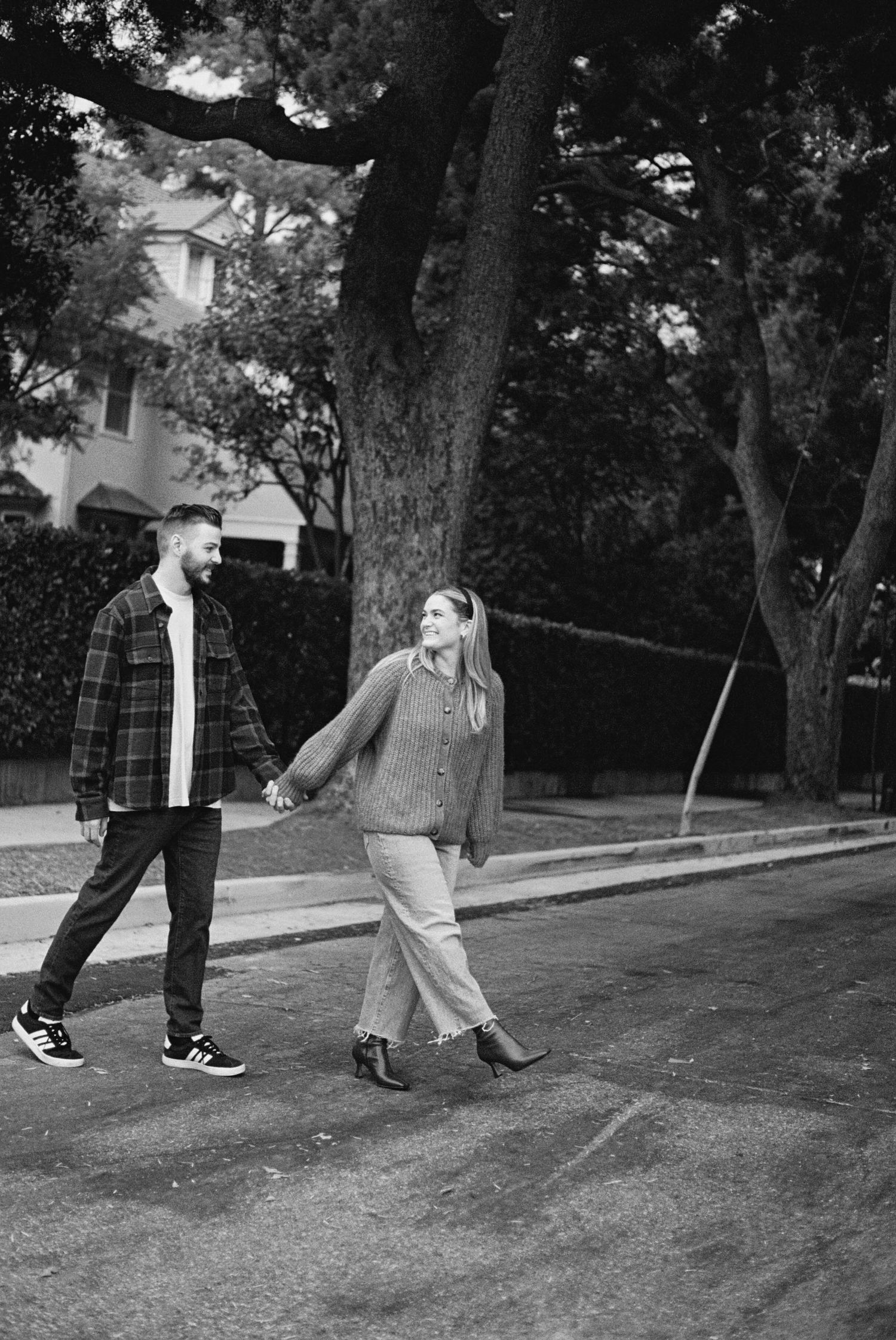 A casual black and white photo of a couple taking a playful stroll down a tree-lined street.