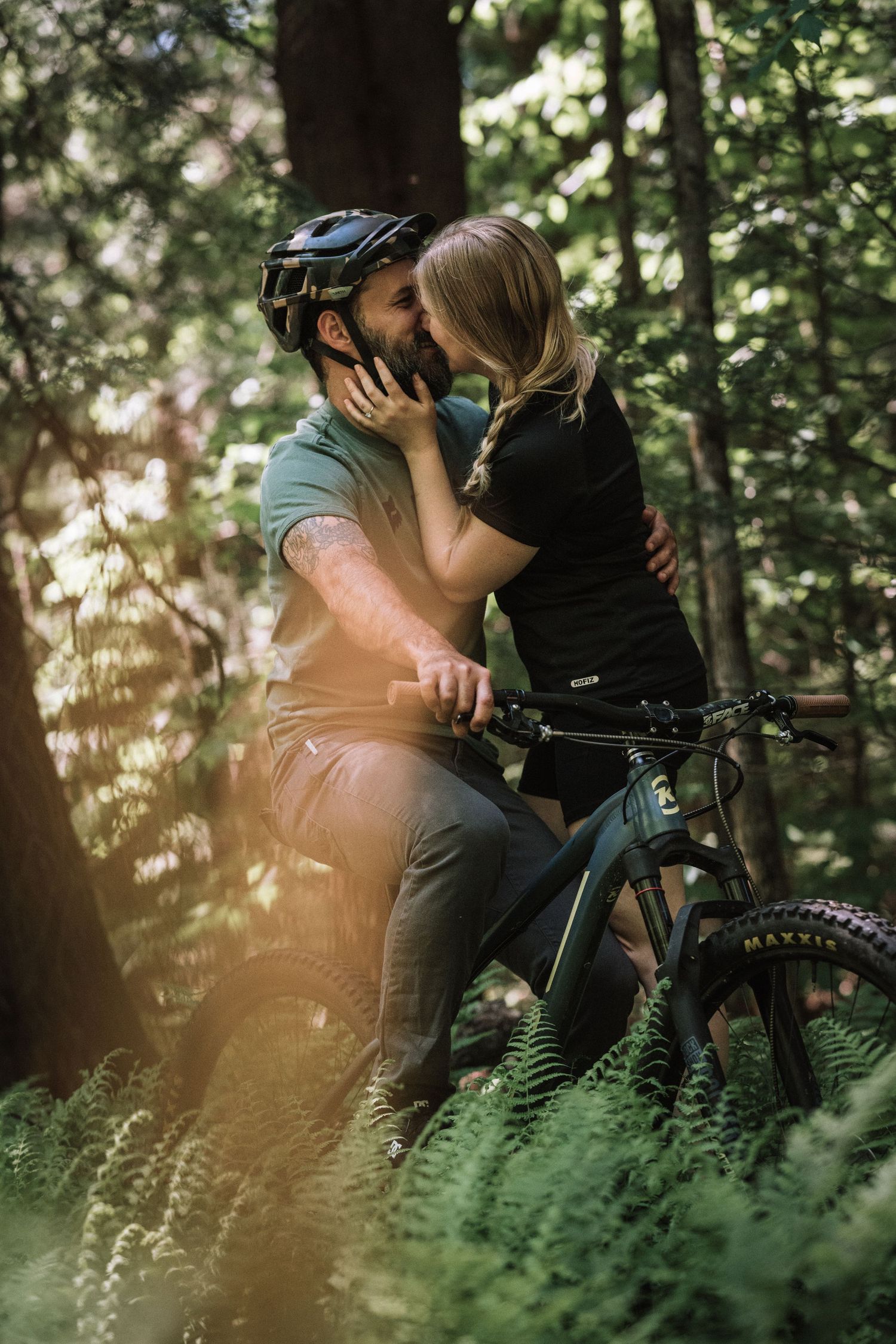 Couple sharing a romantic kiss in a sunlit forest with a mountain bike in the foreground.