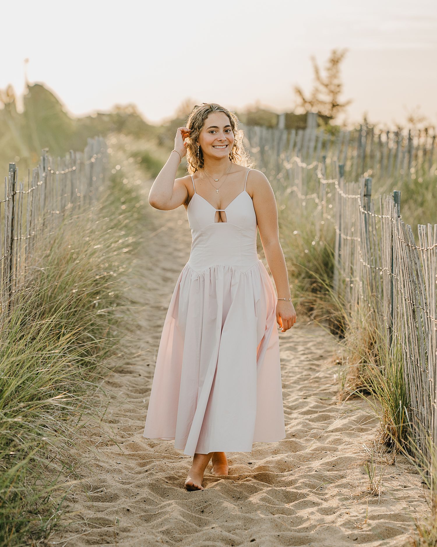 A senior girl in a flowing pale pink dress poses gracefully on sandy dunes during a sunset beach photoshoot in New Hampshire