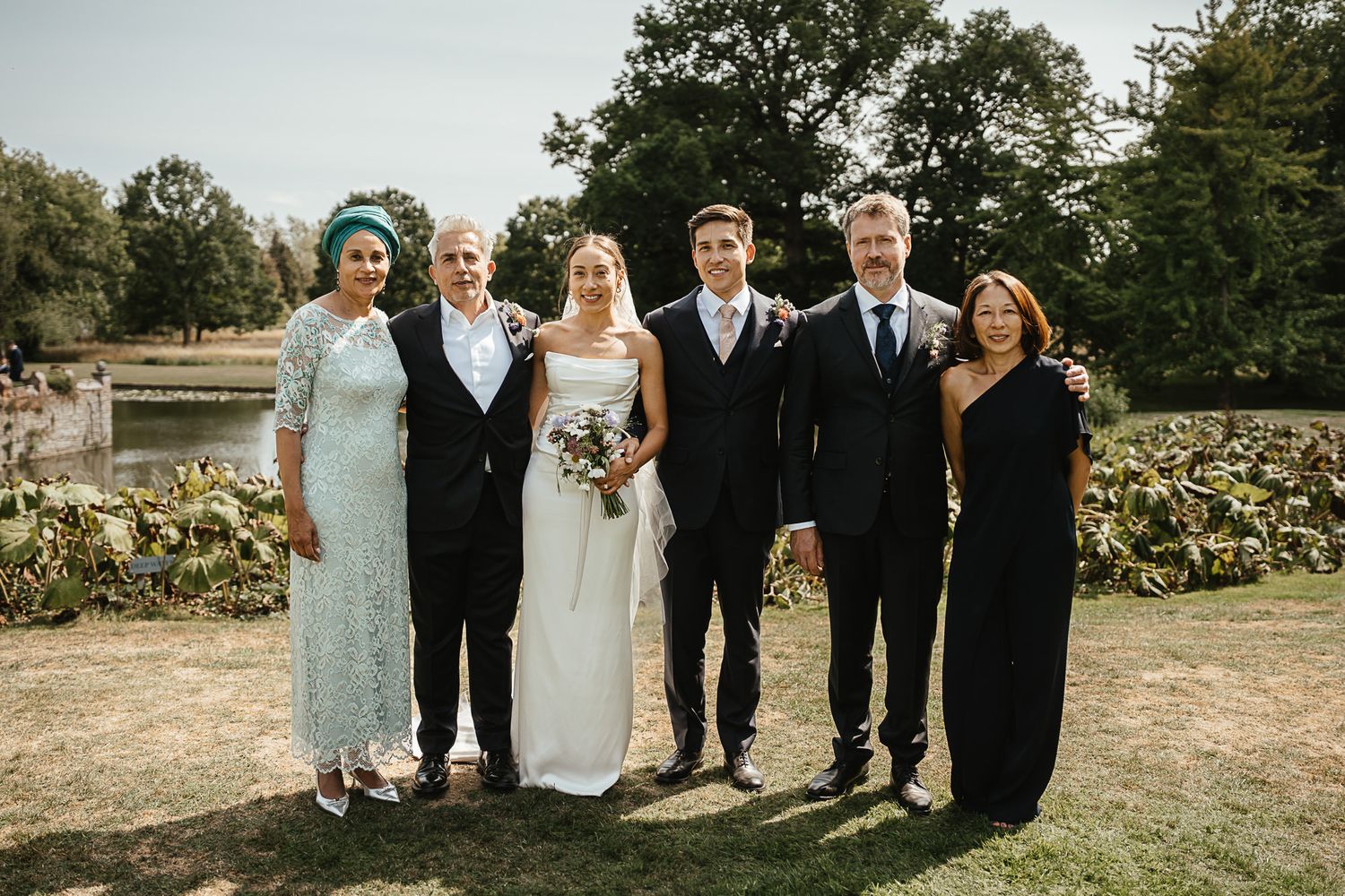 A bridal party poses together on a grass lawn outside a stately brick building, wearing dark green dresses and floral prints.