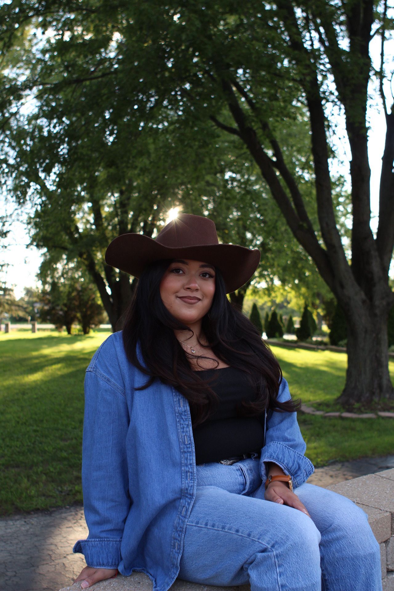 Person in denim jacket and brown cowboy hat sits outdoors under green trees.