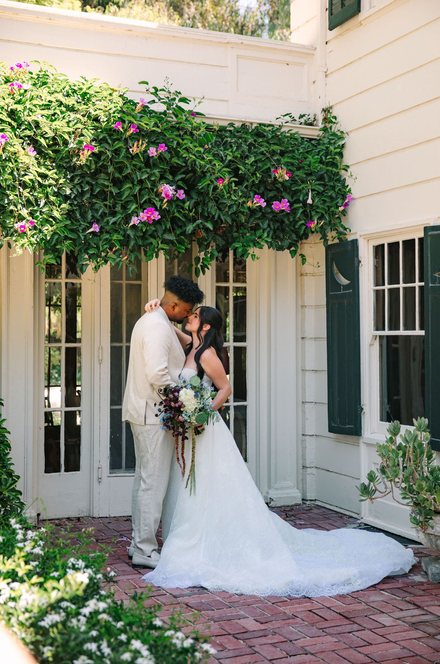A romantic wedding couple embraces in front of a white house with pink climbing roses and green shutters.