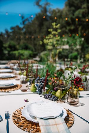 A beautifully set outdoor dining table with white linens, wooden chargers, and delicate purple floral accents stretches into the distance.