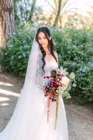 A bride in a flowing white gown holds a vibrant burgundy and white bouquet in a garden setting.