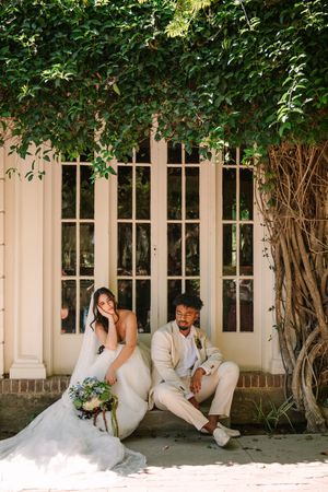 Wedding couple sitting together on garden patio with French doors and vines, sharing a laugh in soft sunlight.