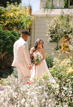 A couple in light colored wedding attire stroll through a sunlit garden with blooming flowers.