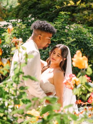A romantic wedding couple embraces among blooming orange and peach colored flowers in a sunlit garden.