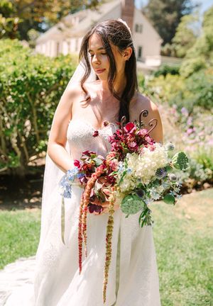 A bride in a white dress holds a beautiful cascading bouquet of burgundy and white flowers in a sunny garden.