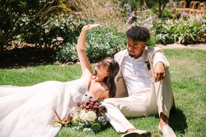 Couple in white attire lounging on sunny grass during outdoor wedding celebration.