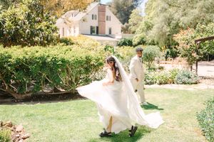 A bride in a flowing wedding dress twirls gracefully in an outdoor garden setting with sunlit trees.