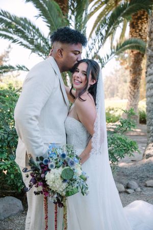 Romantic wedding couple embraces among palm trees in a desert garden setting with elegant white and burgundy floral bouquet.
